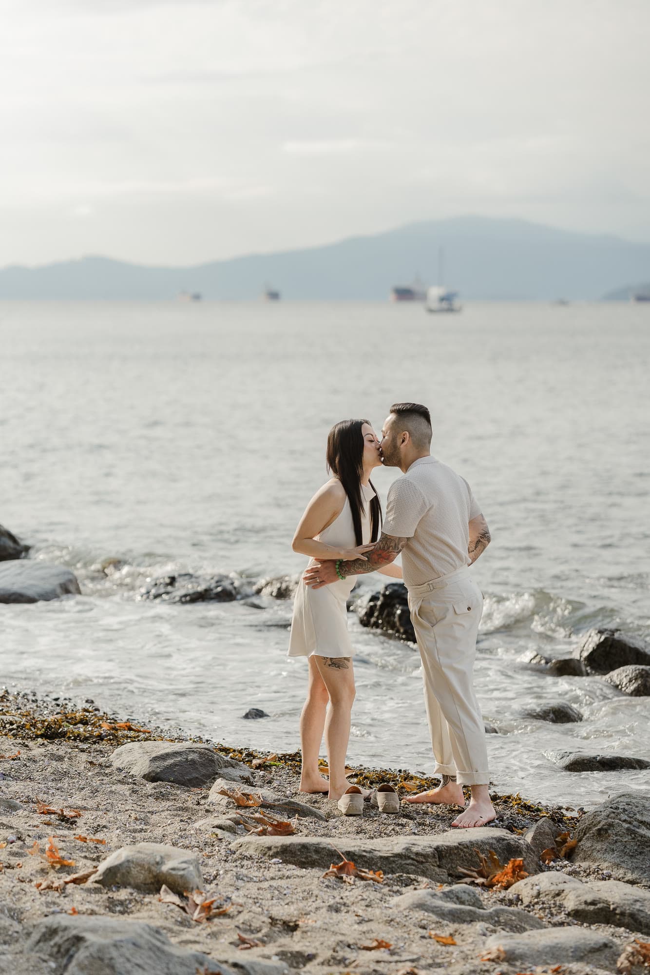 Alana and Colin sharing a kiss at the water’s edge, with boats and mountains behind them.